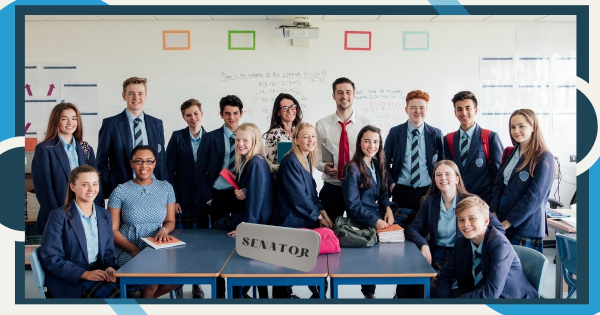 A group of students and teachers posing in a classroom, dressed in formal uniforms and business attire, suggesting a role-playing or simulation activity. The word 'SENATOR' is displayed prominently on a sign at the front of the desk. The whiteboard in the background is covered with equations and notes, indicating an educational setting.