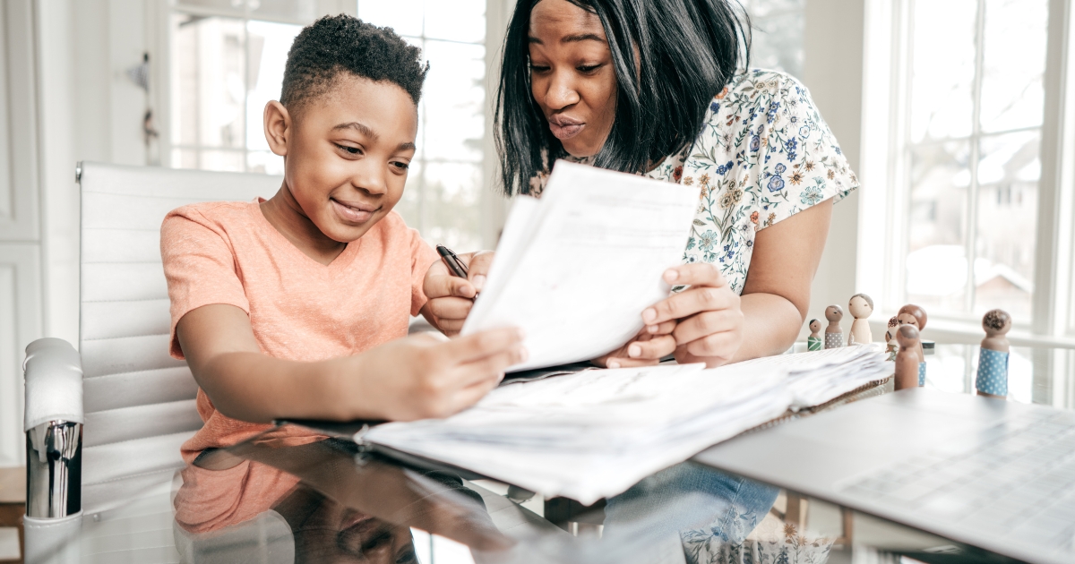 A mother and her young son engaged in a homeschooling session at a table. The boy, wearing an orange shirt, smiles as he looks at a worksheet, while the mother, in a floral blouse, explains something with a pen in hand. The setting is a bright room with large windows and wooden figurines on the table, creating a warm and supportive learning environment.