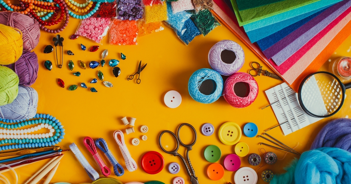 An assortment of colorful DIY art and craft supplies spread on a vibrant yellow background, including yarn, buttons, beads, felt sheets, sewing tools, ribbons, and a magnifying glass, creating a creative and inspiring workspace.