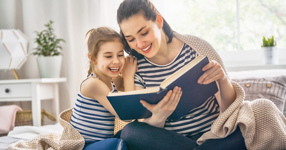 A smiling mother and her young daughter sitting on a cozy couch, reading a book together while wrapped in a blanket, with a bright and airy room in the background.