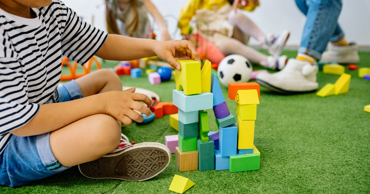 A child in a striped shirt and denim shorts building a colorful tower with wooden blocks on a green carpet, surrounded by other children playing with toys in the background.