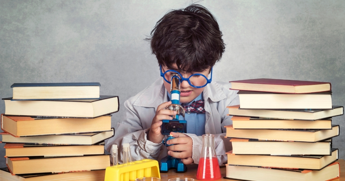 A young boy dressed as a scientist, wearing large blue glasses and a lab coat, intently looking through a microscope. He is surrounded by stacks of books and laboratory equipment, including test tubes and beakers filled with colorful liquids, creating a science-themed setup.