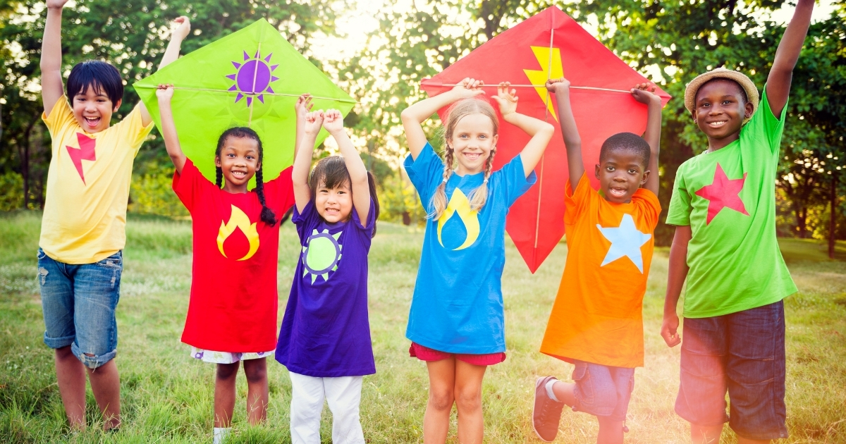 A group of children outdoors wearing colorful shirts with symbols like lightning bolts, flames, and stars, holding kites and smiling, representing fun and active play."