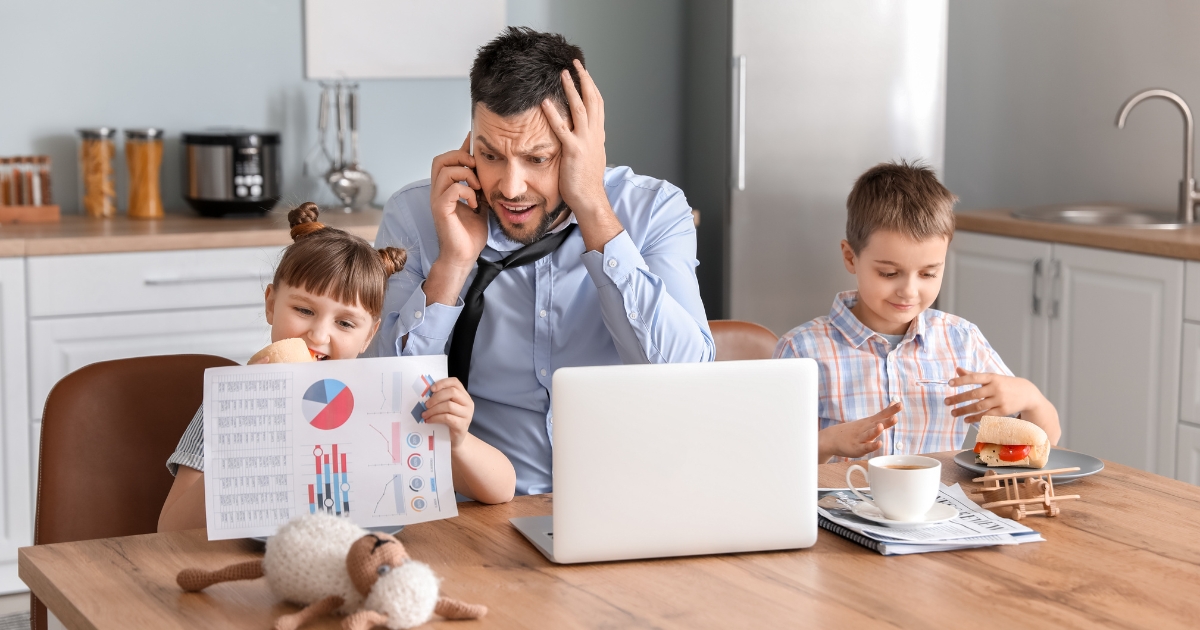 A stressed father in a suit sitting at a kitchen table, talking on the phone while working on a laptop, with two children beside him; one holding a colorful chart and the other eating a sandwich, depicting the challenges of balancing work and homeschooling.