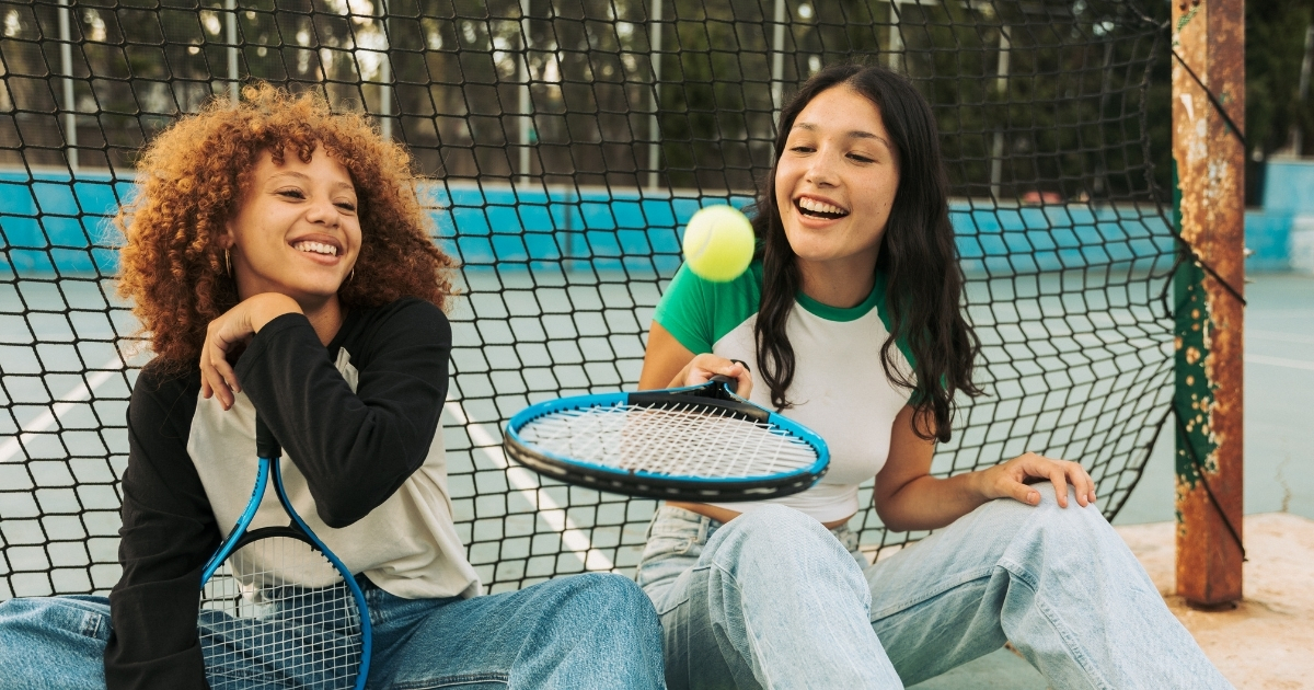 Two teenage girls sitting and laughing on a tennis court, holding rackets and playing with a tennis ball, showcasing fun and active bonding during sports activities.