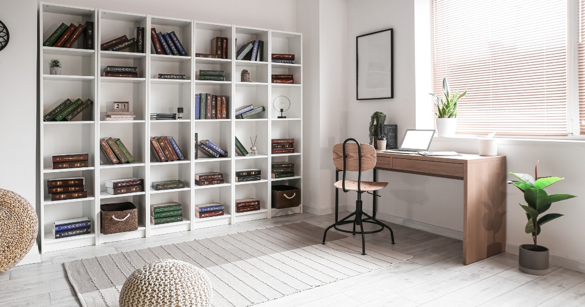 Bright and organized homeschool or study room with a white bookshelf filled with colorful books, a wooden desk with a laptop and potted plants, a woven pouf, and a cozy beige rug on light wood flooring.