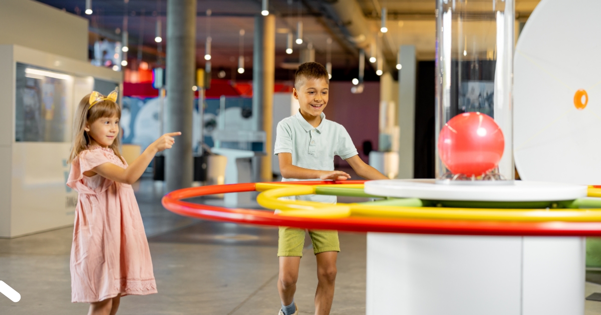 Two children enjoying an interactive science exhibit at a museum, with a young boy smiling while operating colorful rings and a young girl pointing at a red ball floating in a clear tube.