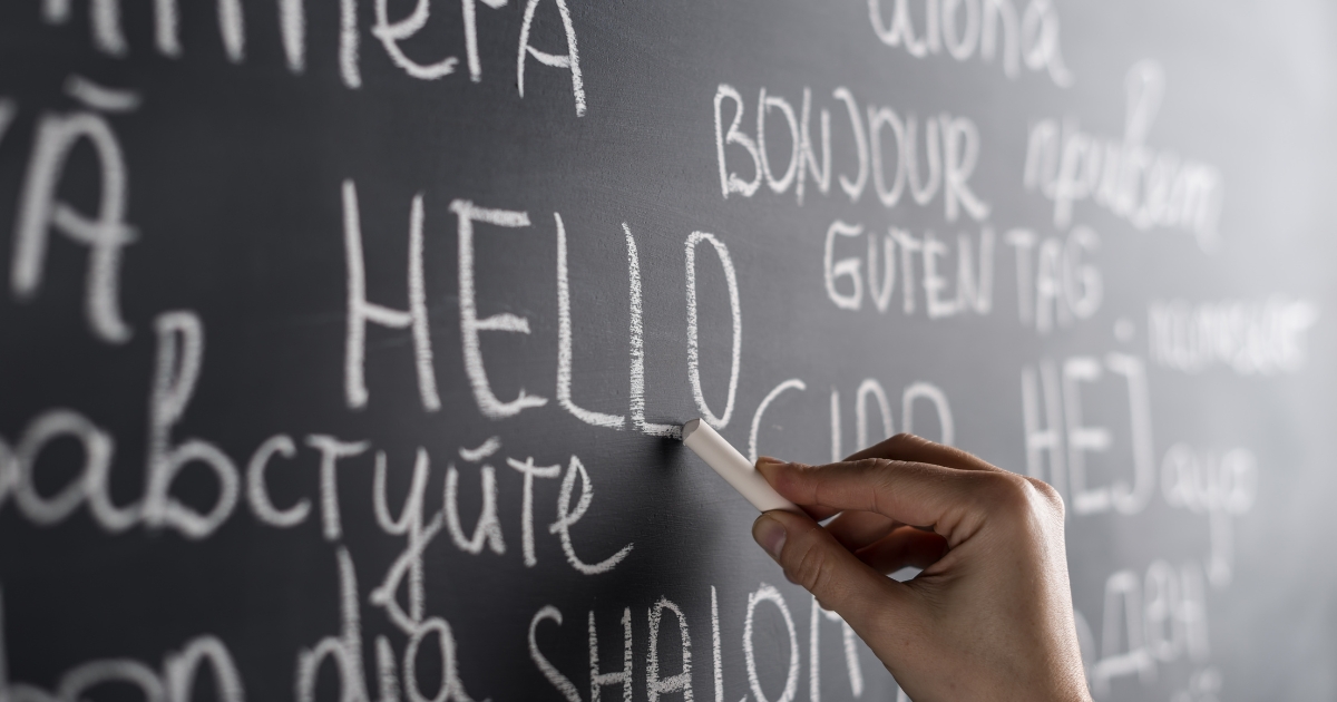 Close-up of a hand writing the word 'HELLO' in chalk on a blackboard, surrounded by greetings in multiple languages such as 'Bonjour,' 'Guten Tag,' and 'Ciao,' symbolizing multilingual education and language learning.