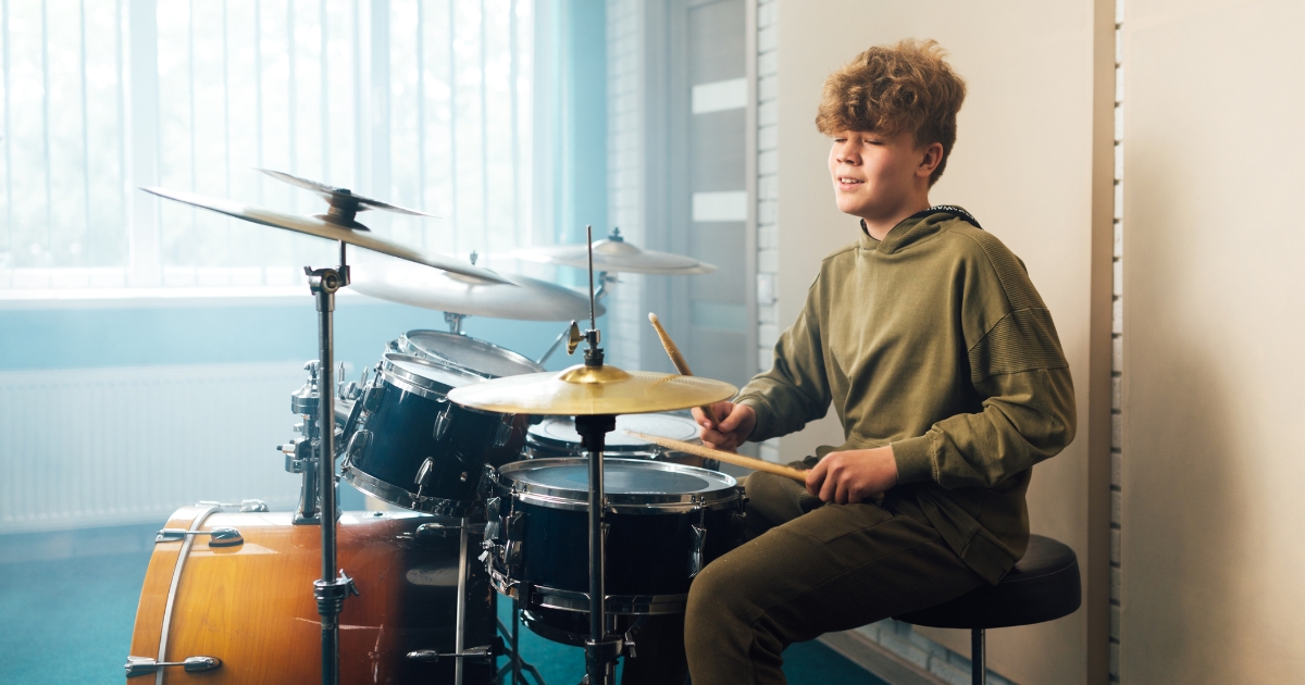 Teenager playing a drum set in a brightly lit room, with natural light streaming through large windows, creating a focused and creative atmosphere.