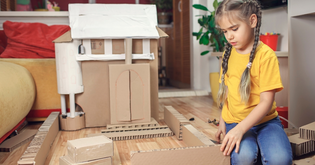 A young girl with braided hair wearing a yellow shirt and jeans is sitting on the floor building a model house out of cardboard. The house features detailed walls, a roof, and windows, set in a cozy indoor space.