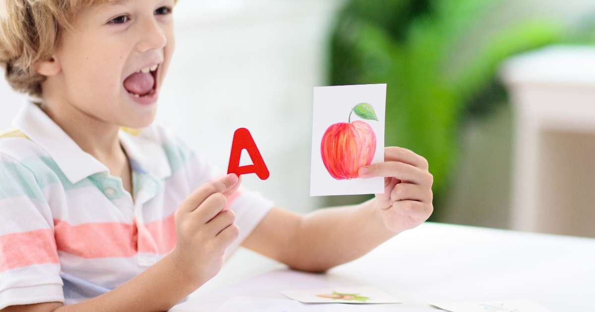 A young child excitedly learning letters and words, holding a red letter 'A' in one hand and a flashcard with a drawing of an apple in the other, sitting at a table with additional cards.