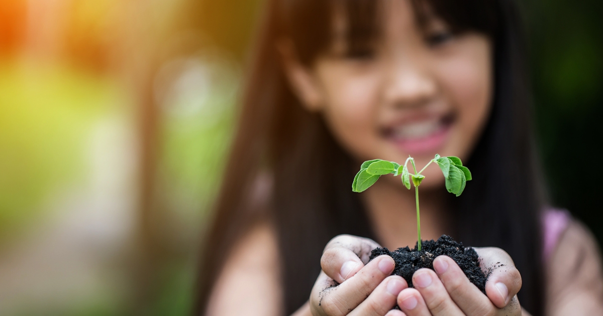 A smiling child holding soil with a small green seedling, symbolizing growth, learning, and hands-on biology experiments in a homeschool setting.