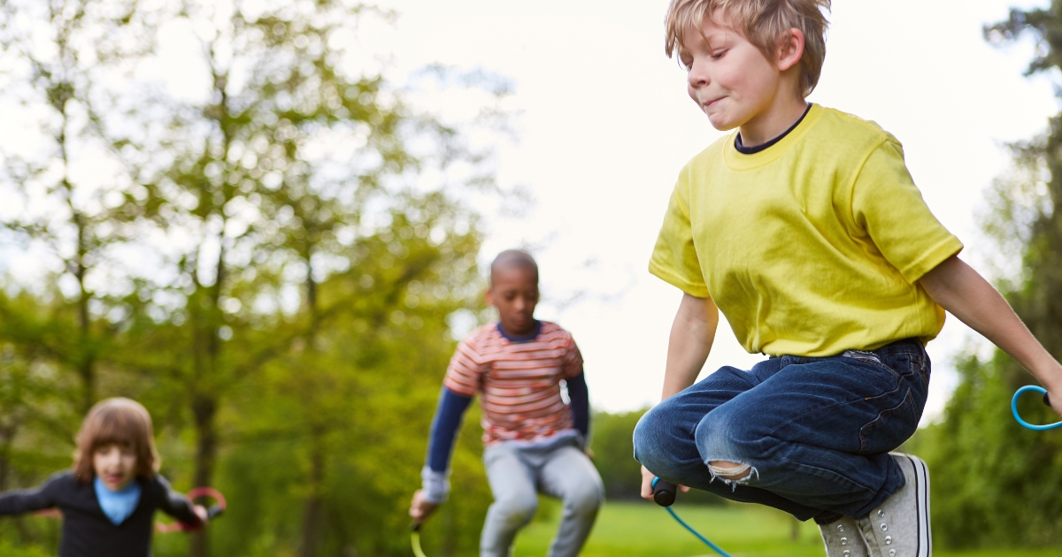 Three children enjoying an outdoor activity, jumping rope in a lush green park, showcasing fun and active fitness during a homeschool gym class.