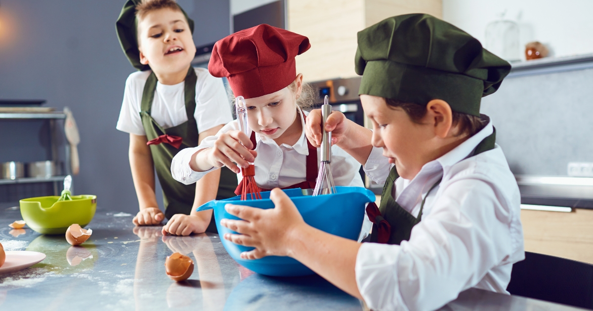 Three children wearing colorful chef hats and aprons mixing ingredients in a bowl during a fun cooking activity, demonstrating hands-on learning and math skills in a homeschool environment.