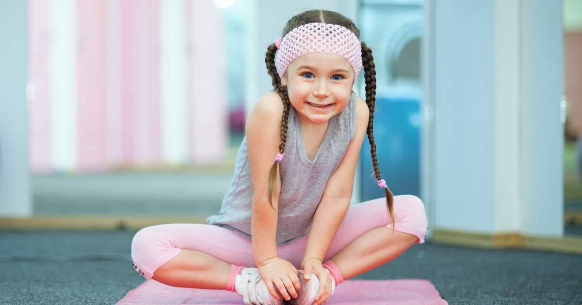 Young girl with braided hair and a pink headband smiling while sitting cross-legged on a yoga mat in a brightly lit fitness studio, promoting physical activity and wellness for children.