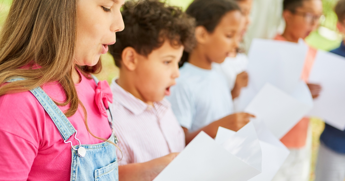 A group of children standing outdoors, holding sheets of paper and singing together in a chorus. The focus is on a girl in a pink shirt and denim overalls, with other children visible in the background, creating a vibrant and collaborative scene.