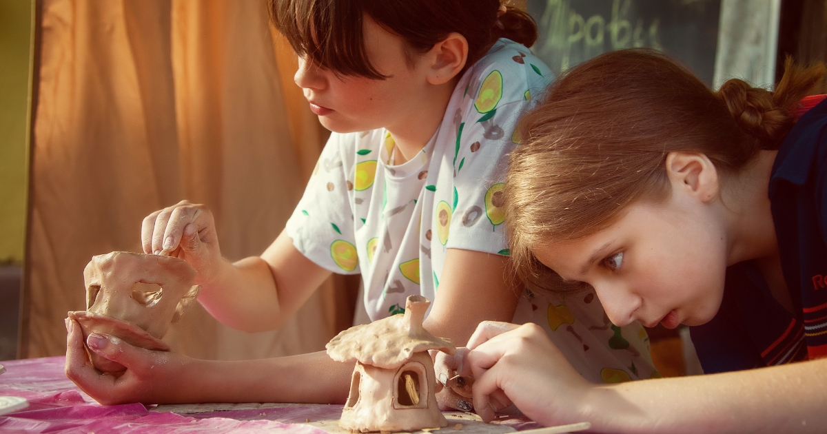 Two children deeply focused on crafting clay sculptures at a table. One child is shaping a hollow object, while the other is adding details to a small clay house. The warm lighting and casual setting create a creative and hands-on atmosphere.