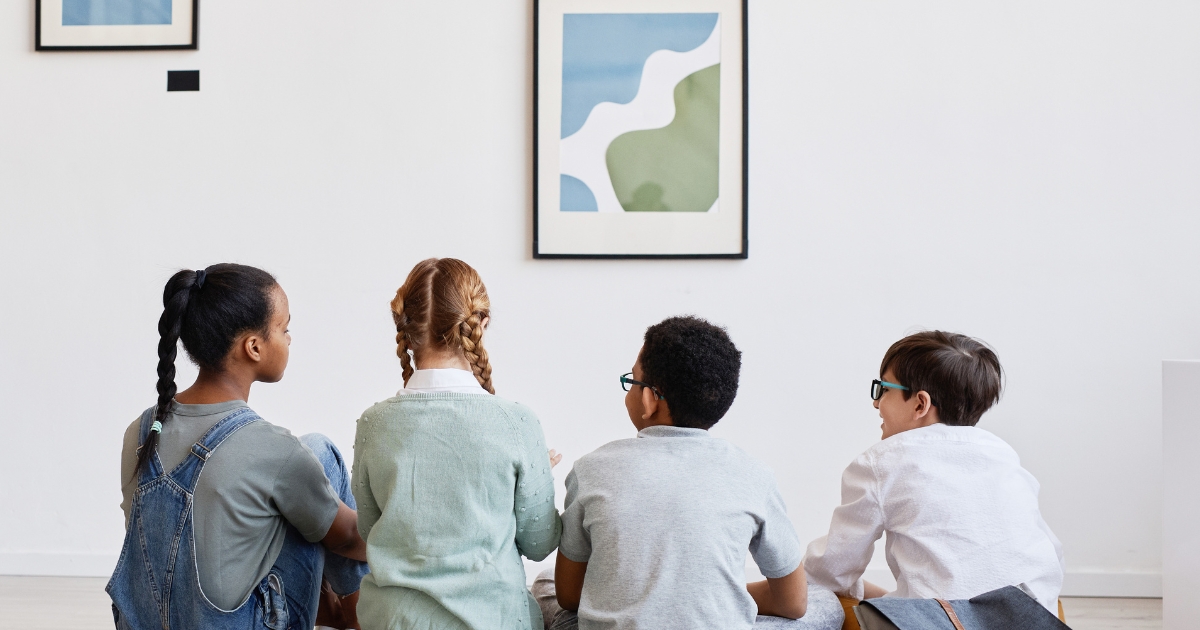 Four children sitting on the floor in an art gallery, viewed from behind, as they observe a framed abstract painting on a white wall.