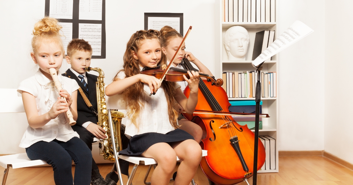 A group of children in a music class playing various instruments, including a recorder, saxophone, violin, and cello. They are seated in a bright room with sheet music on the wall and bookshelves in the background, creating a classroom-like environment.