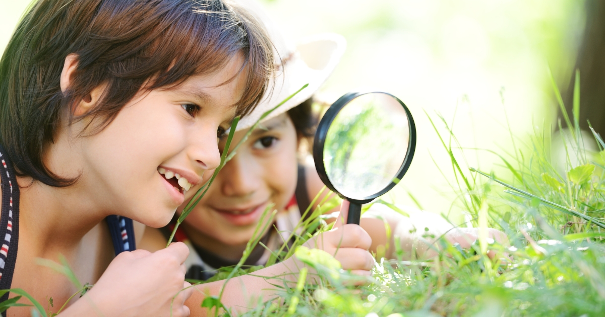 Two children lying on the grass outdoors, smiling and using a magnifying glass to observe small details in the greenery. The scene is bright and sunny, reflecting an engaging and curious exploration of nature.