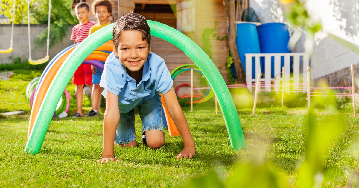 A young boy crawling through a colorful pool noodle tunnel in a backyard obstacle course, with other children watching in the background, enjoying an outdoor activity.