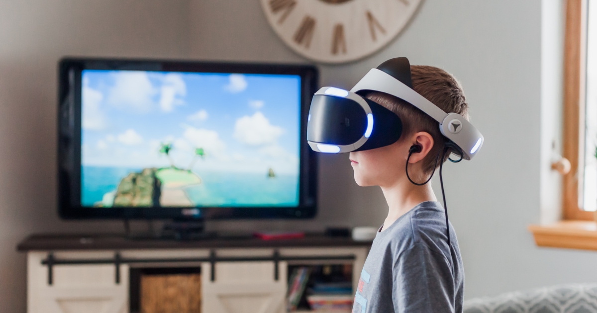 A young boy wearing a virtual reality headset, immersed in a digital experience, with a TV screen in the background displaying a tropical island scene in a home setting.