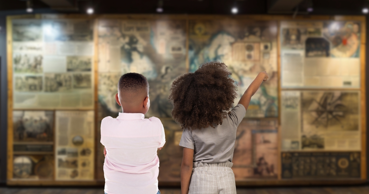 Two children standing in front of a large historical map display in a museum, with one child pointing toward the map, engaged in exploring history.