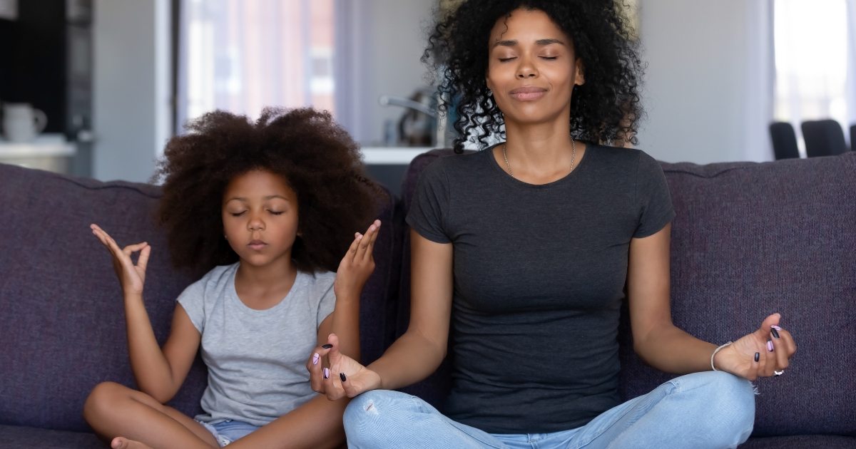 A mother and her young daughter sitting cross-legged on a sofa, practicing mindfulness and meditation with their eyes closed and hands in a meditative gesture, in a serene home setting