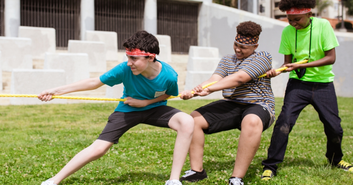 Three teenagers participating in a game of tug-of-war on a grassy field, wearing colorful bandanas and showcasing teamwork and determination during an outdoor activity.