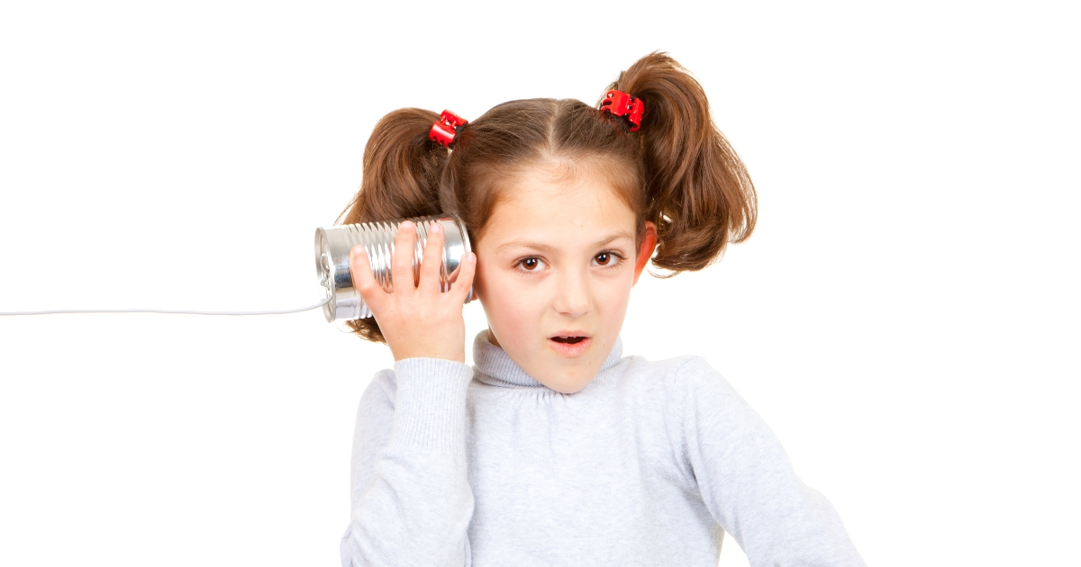 A young girl with pigtails and red hair clips holding a tin can connected to a string, pretending to listen, demonstrating a classic sound experiment.