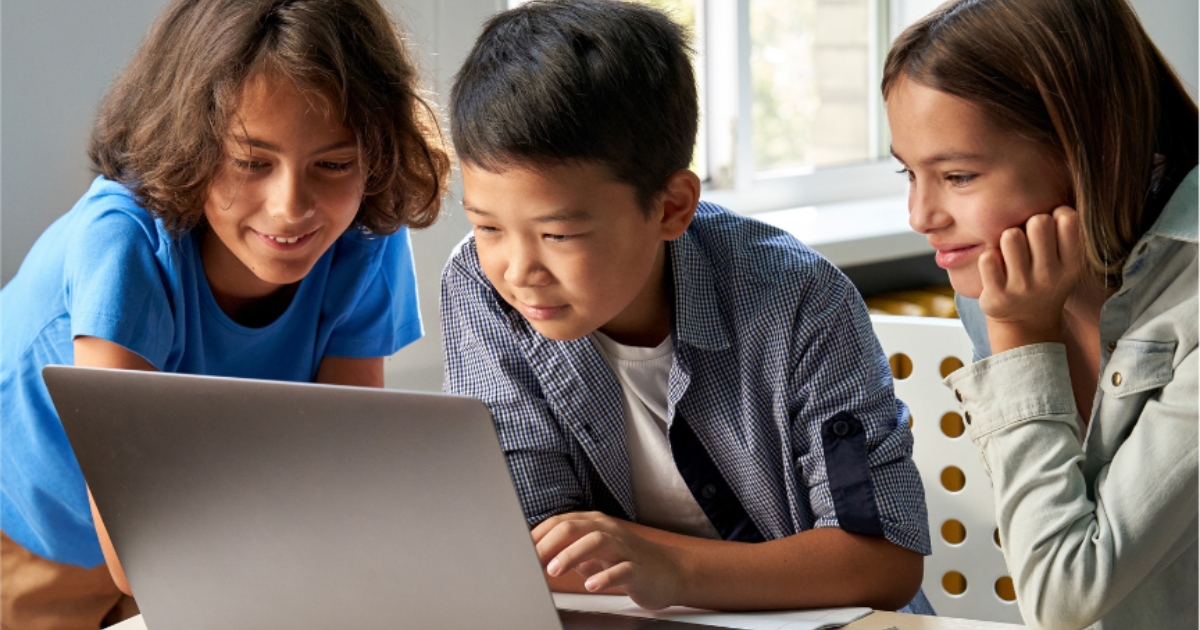 Three children smiling and collaborating while looking at a laptop screen in a brightly lit room, seated at a table.