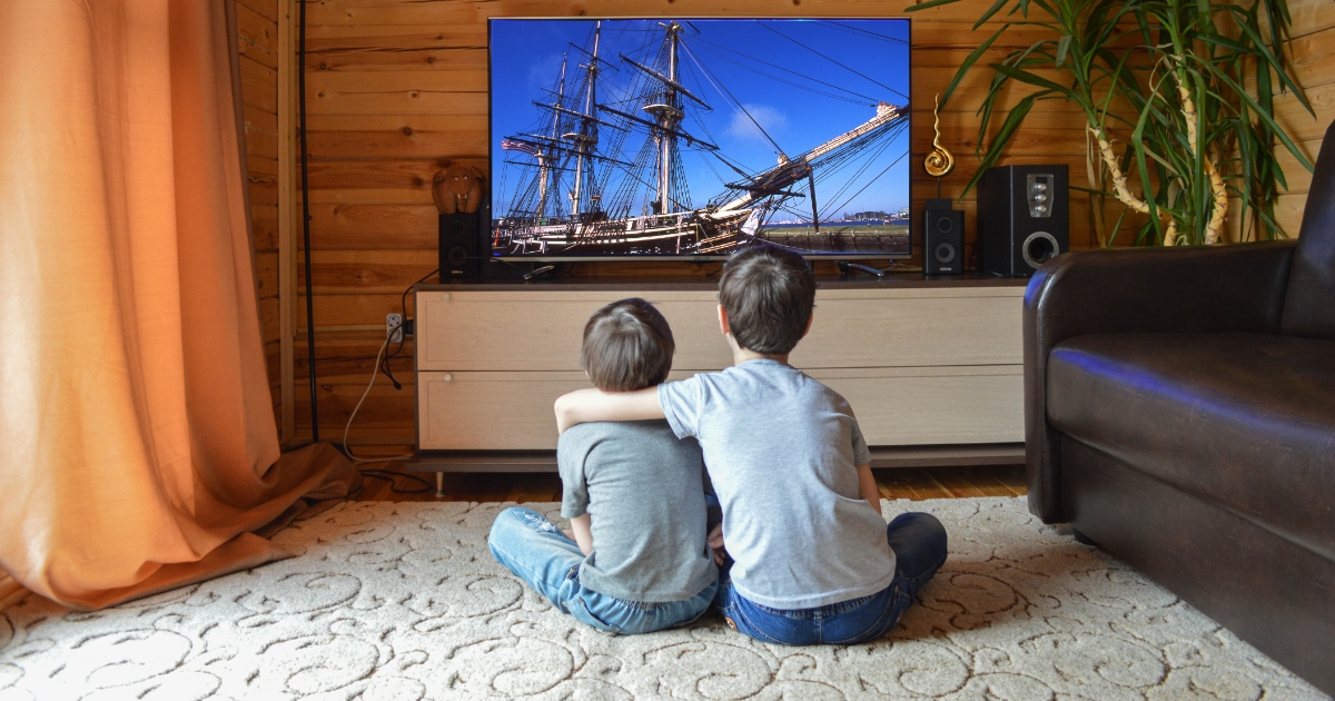 Two young children sitting on the floor in a cozy wooden living room, watching a historical ship on a large TV screen, with one child’s arm around the other.