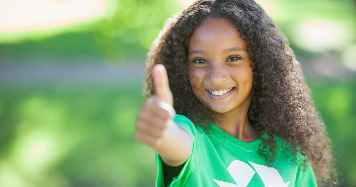 A cheerful young girl outdoors giving a thumbs-up, wearing a green shirt with a recycling symbol, symbolizing positivity and environmental awareness. The background is lush and blurred, creating a bright and natural atmosphere.