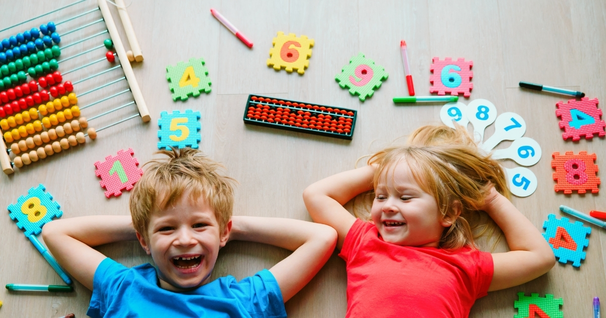 Two smiling children lying on the floor surrounded by colorful numbers, abacuses, and math learning tools, symbolizing playful and engaging math activities for homeschool education.