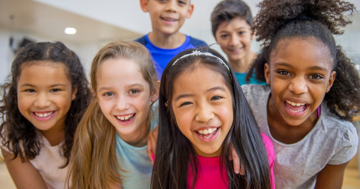 A diverse group of six smiling children standing close together indoors, looking cheerful and excited in a brightly lit room.