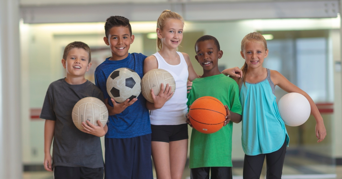 A diverse group of five smiling children standing indoors, each holding a sports ball including a soccer ball, volleyball, basketball, and white foam ball, symbolizing teamwork and a variety of sports activities.