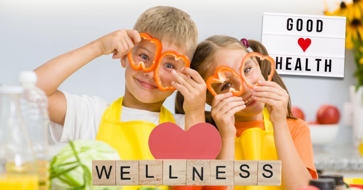 Two children wearing aprons holding red bell pepper slices shaped like hearts over their eyes, smiling in a kitchen setting, with the words 'Good Health' and 'Wellness' displayed, promoting healthy eating and wellness education.