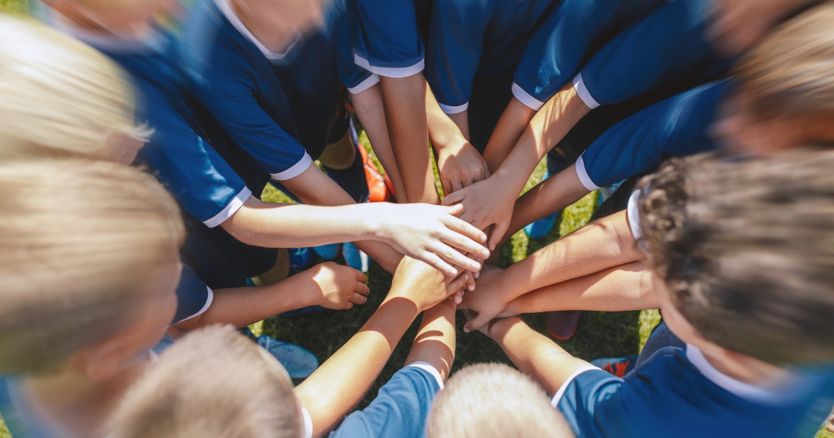 A group of children in blue sports uniforms placing their hands together in a huddle, symbolizing teamwork and unity on a grassy field.