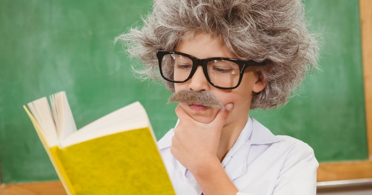 A young child dressed as Albert Einstein, wearing a white lab coat, oversized glasses, a fake mustache, and a wild gray wig, thoughtfully reading a yellow book in front of a green chalkboard. A fun and creative approach to learning through role-playing.