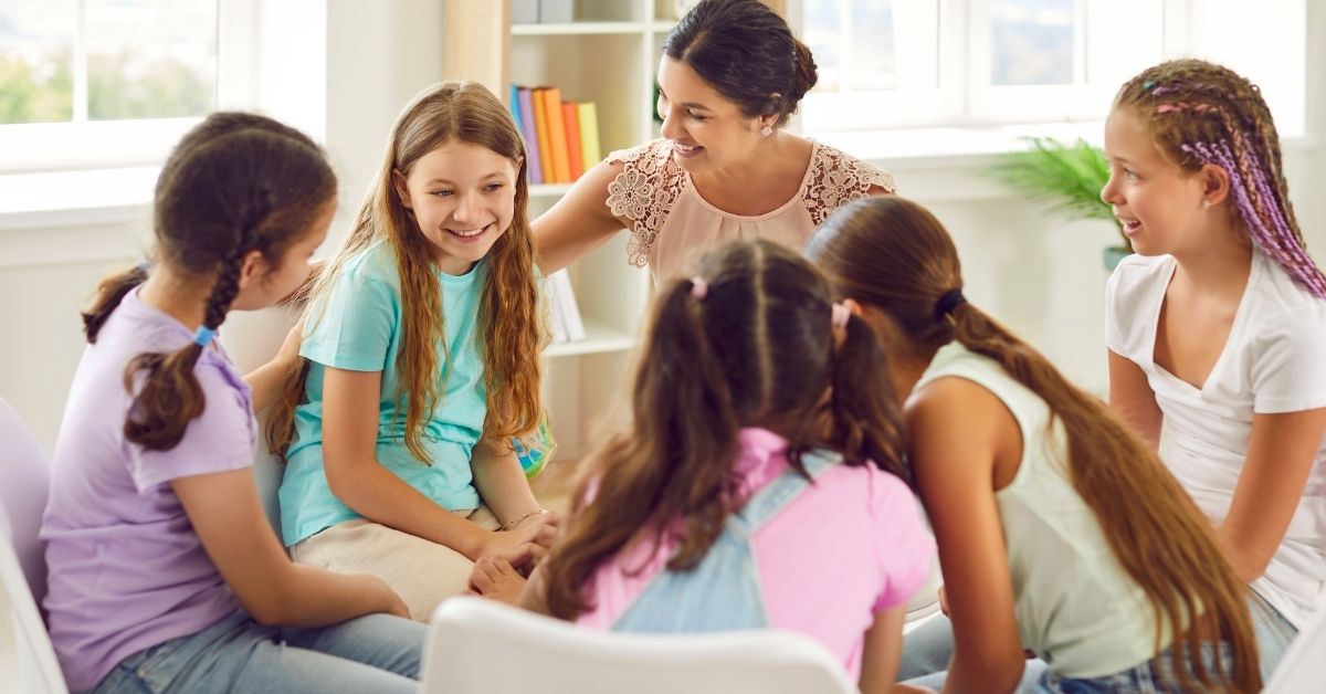 A group of young girls sitting in a circle engaging in a discussion with a smiling female teacher or mentor. The children appear happy and engaged, while the teacher encourages participation. The setting is a bright and welcoming indoor learning environment with bookshelves and natural light in the background.