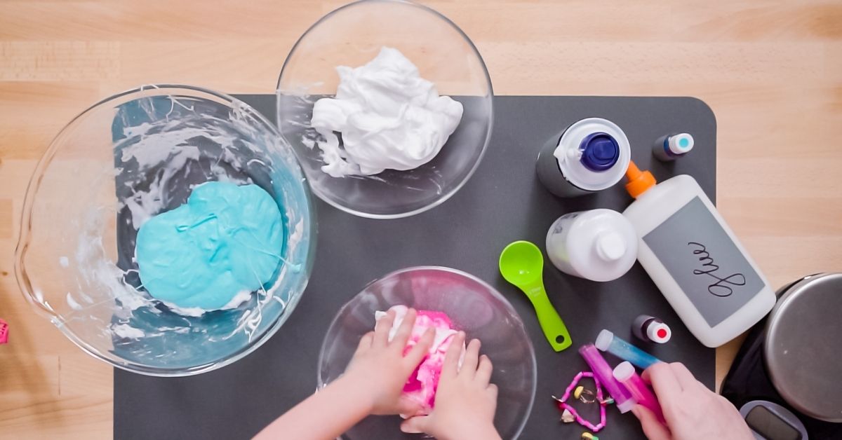 A top-down view of a child's hands kneading pink slime in a bowl, surrounded by various slime-making ingredients and tools, including glue, food coloring, shaving cream, measuring spoons, and mixing bowls containing blue and white mixtures. A fun and colorful hands-on science experiment in progress.