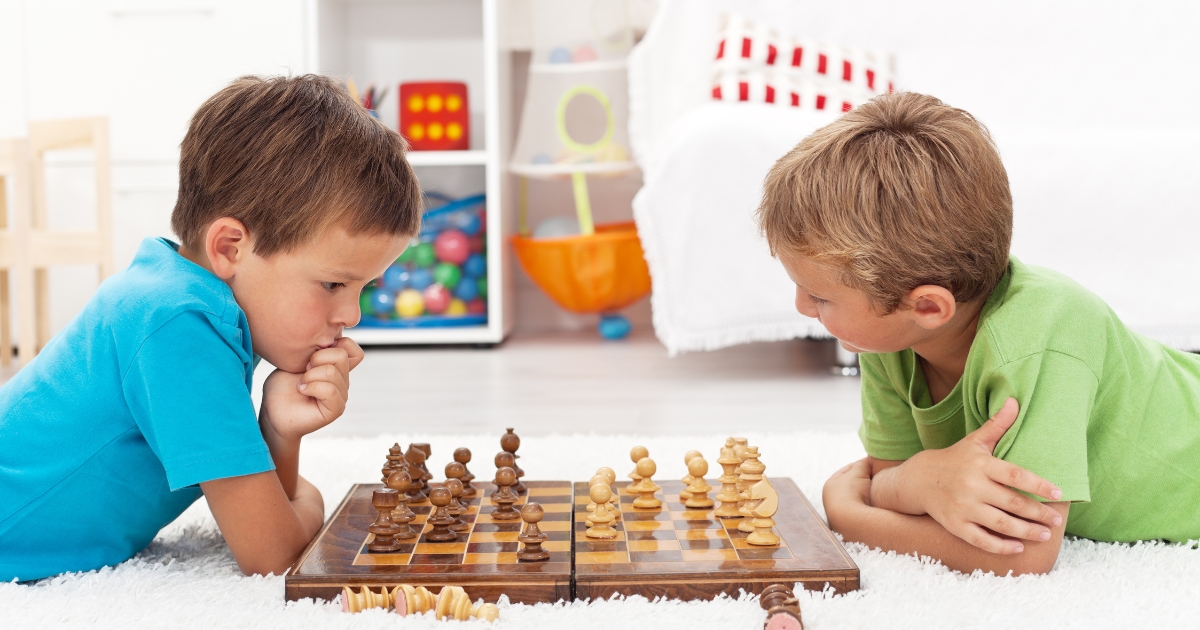 Two young boys playing chess on the floor in a bright playroom, focusing on strategy and decision-making. The image represents critical thinking, problem-solving, and independent learning in childhood development.