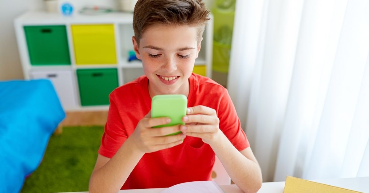 A young boy wearing a red shirt sits at a desk, smiling while looking at a green smartphone. His study materials, including a notebook, are in front of him, and colorful storage cubes decorate the background. A bright and engaging learning environment.