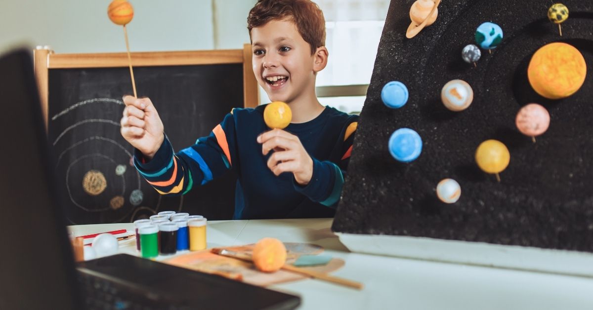 A young boy with a bright smile engages in a hands-on science project, holding a small model of a planet. He is working on a solar system model, with colorful planets attached to a black display board. Behind him, a chalkboard features a hand-drawn solar system, and on the table, there are paint bottles and craft supplies. The scene depicts a creative homeschooling or learning environment.