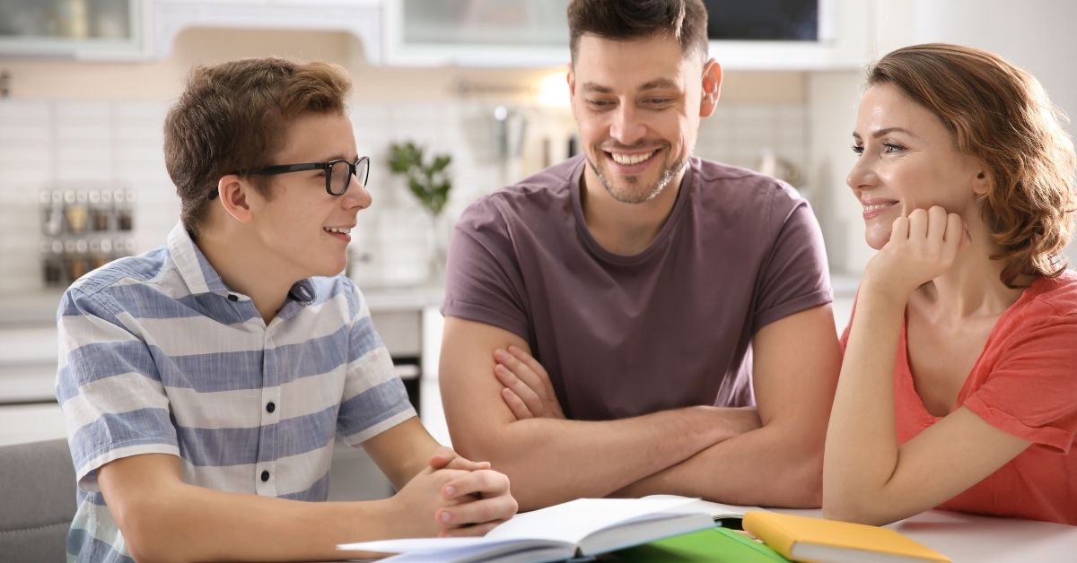 A teenage boy wearing glasses and a striped shirt sits at a table with his parents, smiling and engaged in conversation. Open books and notebooks are spread out on the table, suggesting a homeschooling or study session in a bright and cozy kitchen setting.