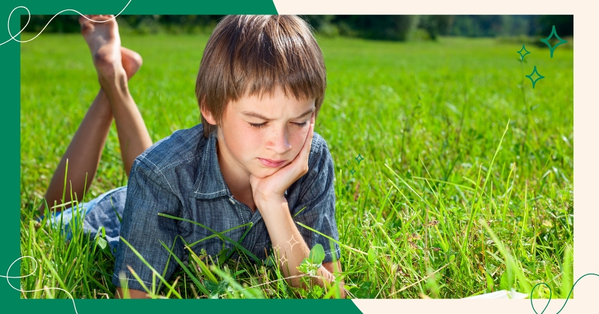 Young boy lying in the grass, deeply focused on reading a book outdoors. The image represents the Charlotte Mason approach to literature, encouraging a love for reading through nature and immersive learning.