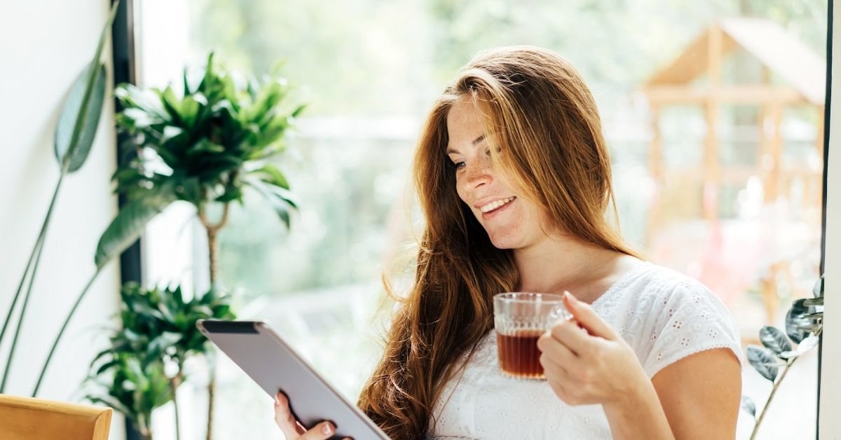 A smiling woman with long red hair, wearing a white lace top, sits near a large window filled with natural light. She holds a glass cup of tea in one hand and a tablet in the other, appearing engaged in what she is reading. Lush green plants surround her, and a blurred wooden play structure is visible outside.