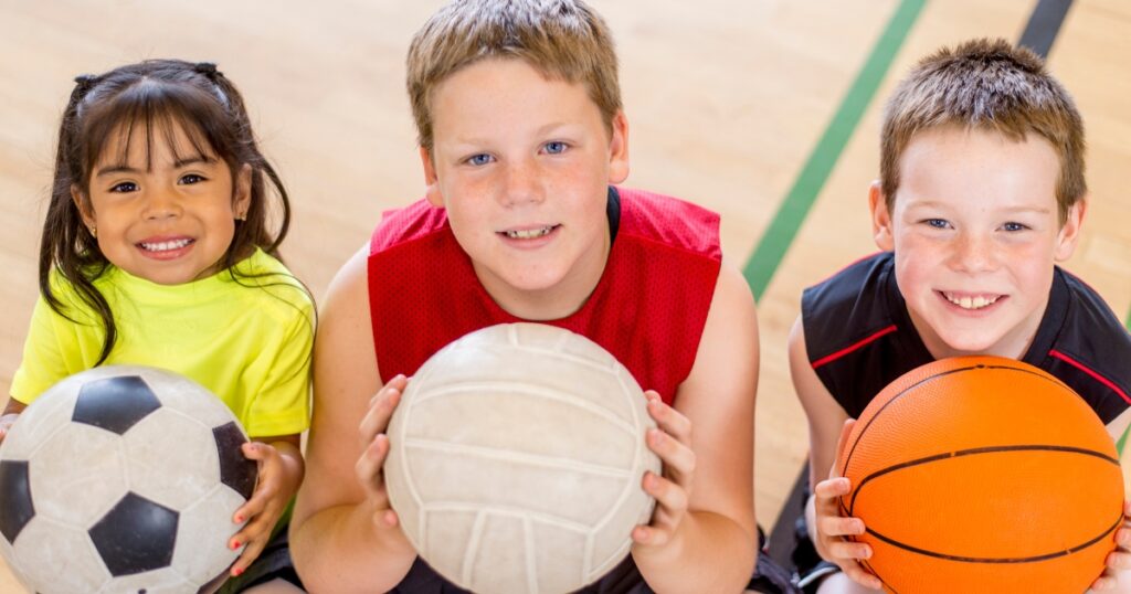 Three smiling children sitting on a gym floor, holding sports balls – a soccer ball, volleyball, and basketball – during a homeschool physical education activity.