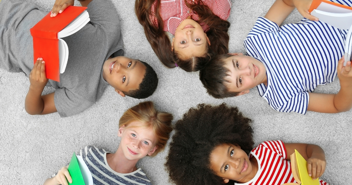 Diverse group of smiling children lying on a soft carpet, holding colorful books and looking up, symbolizing confidence and joy in learning through literature.