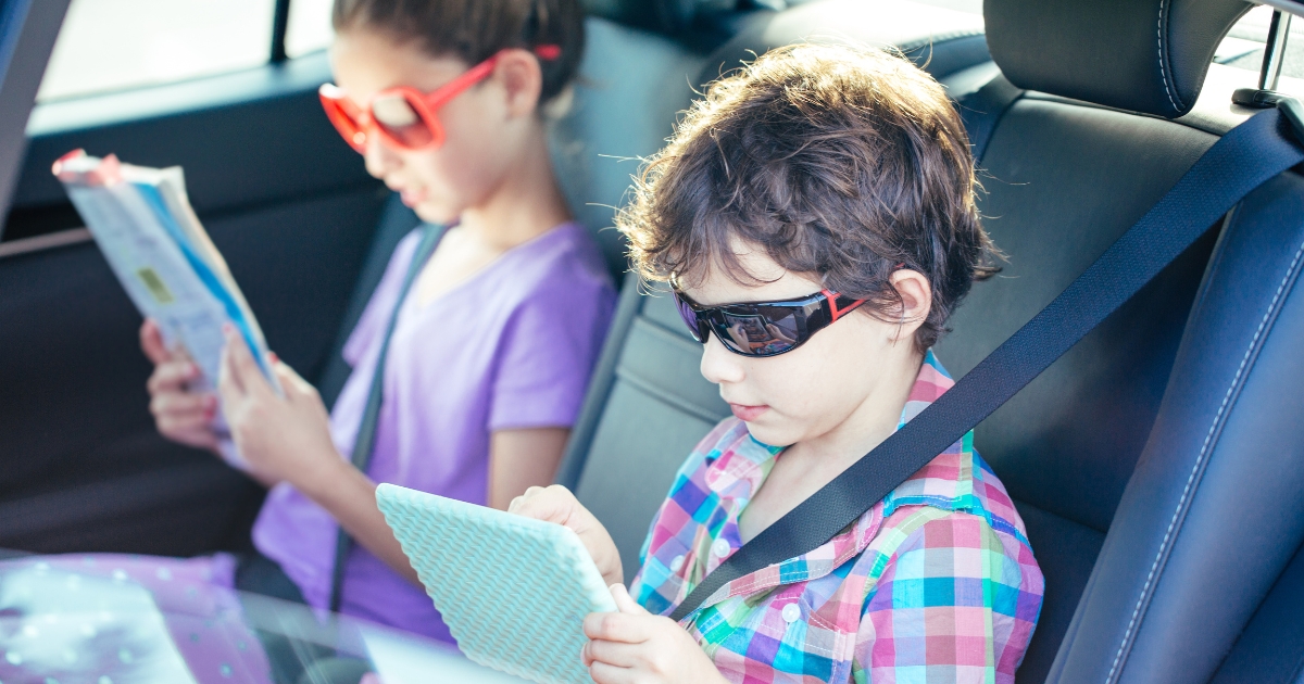 Children homeschooling while traveling, reading a book and using a tablet in the backseat of a car.